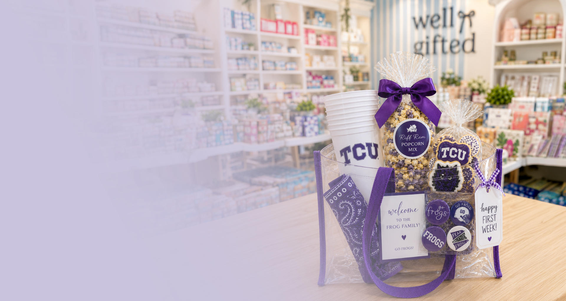 A TCU football-themed gift basket set on a wooden table inside a gift shop, featuring a white cup with 'TCU' printed on it, a bag of gourmet snacks, a purple ribbon-wrapped item, and a 'Welcome to the family' card, with a blurred background showing store shelves and a 'well gifted' sign.